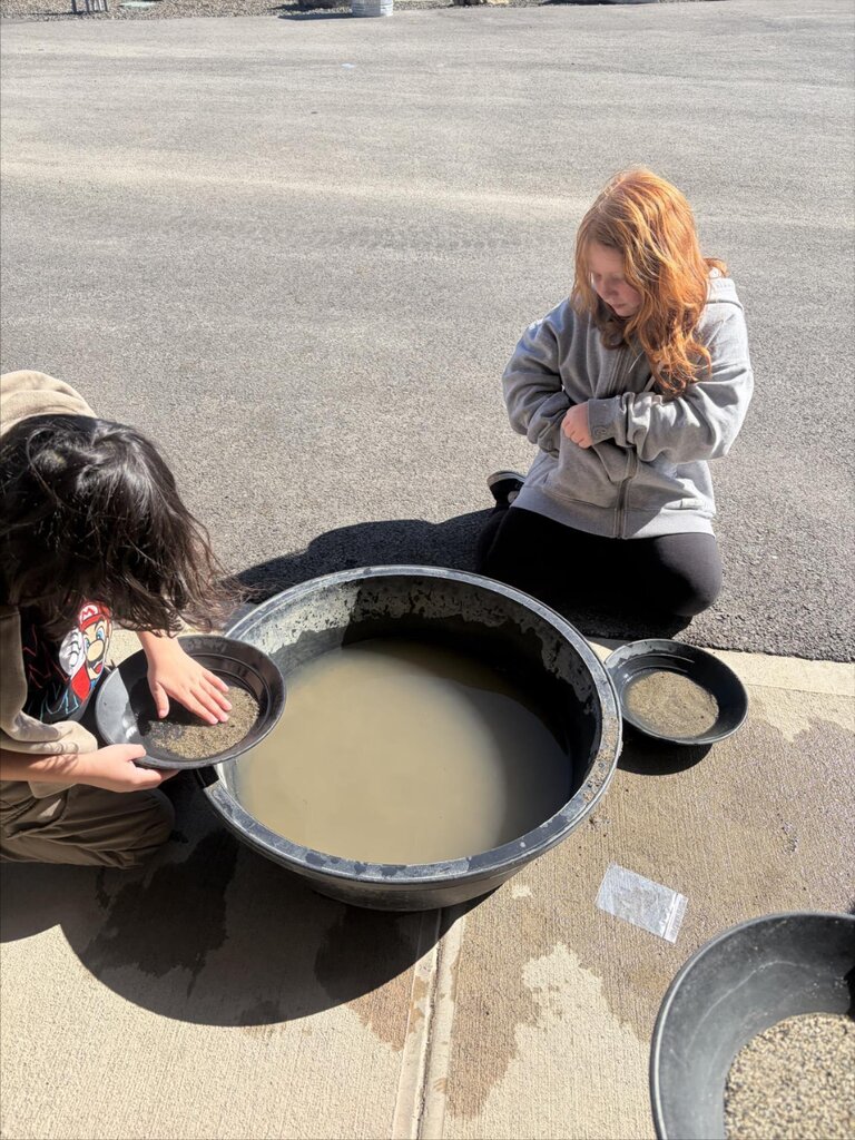 Two individuals kneel by a large basin with water on a paved surface. One stirs water, the other examines a pan.