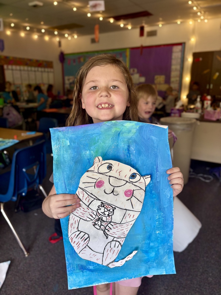 A child displays a watercolor painting of a cat, smiling, in a classroom with desks, chairs, and a trash bin.