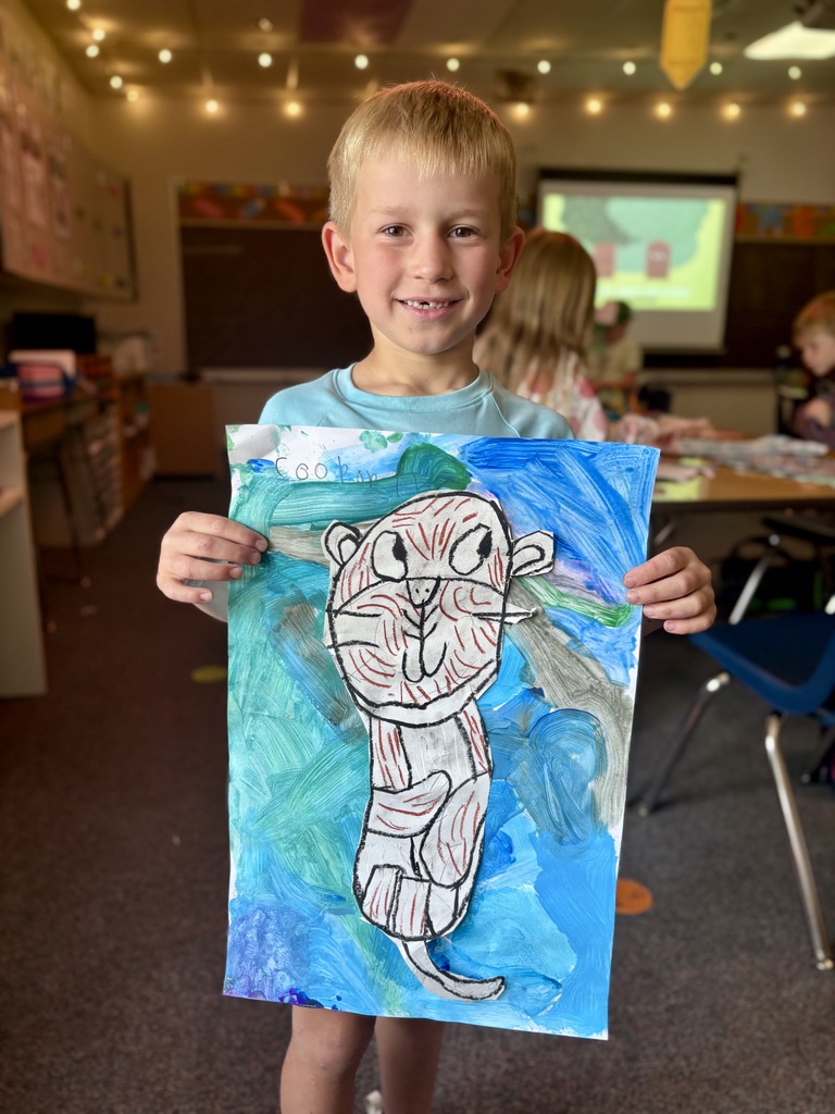 A boy holds a drawing of a tiger in a classroom. Behind him, a projector screen is visible, and children work at tables.