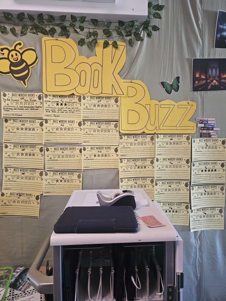 A wall with a yellow sign that reads "Book Buzz" with a bee and a butterfly. Below it are yellow papers. In front is a white desk with a black tablet and papers.