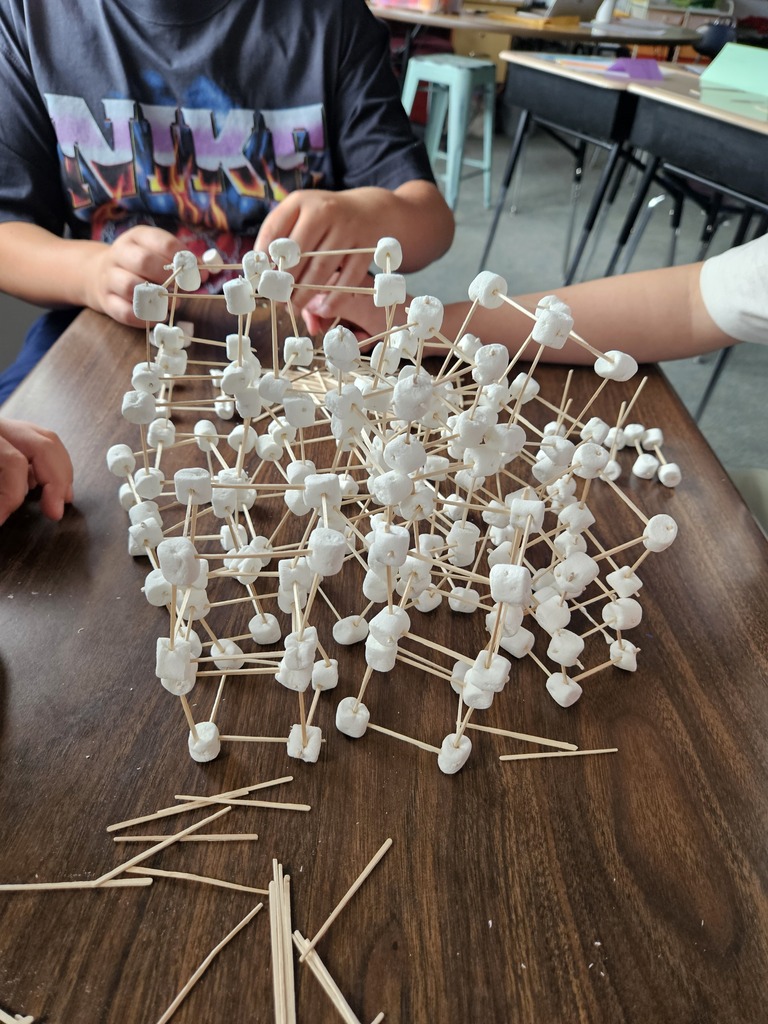Two individuals working on a table with a pile of marshmallows and toothpicks. Background includes desks and chairs.