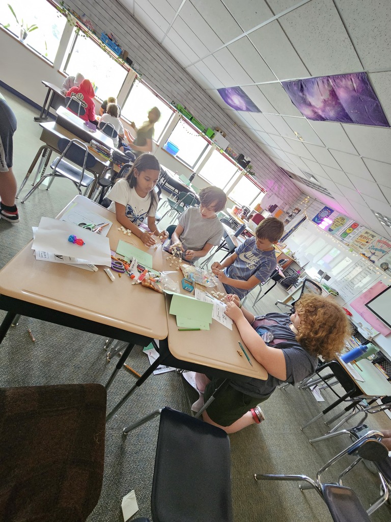 A classroom with children at desks and a teacher assisting them. Tables have art supplies.