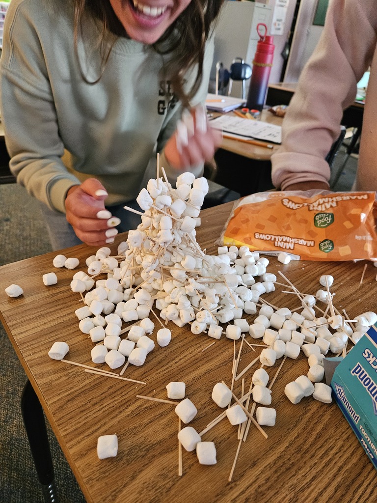 A person uses skewers to arrange marshmallows on a desk. Another person watches. A box of marshmallows and a thermos are nearby.