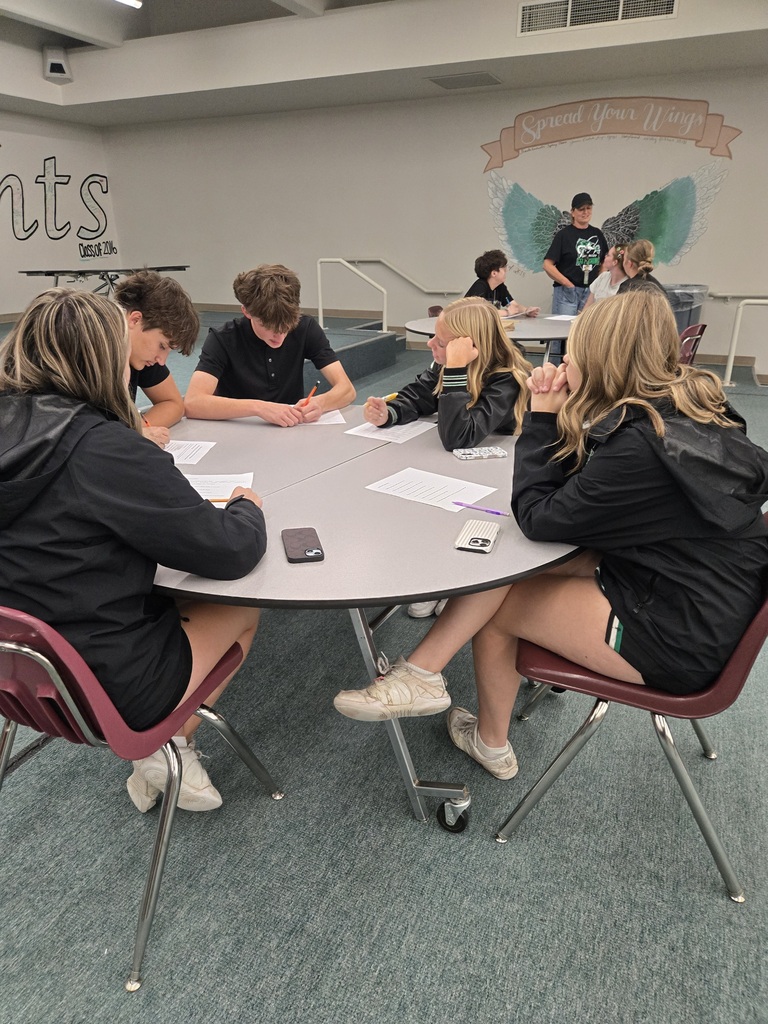 Students seated at a table in a room with a banner, working on papers. A person stands behind them.