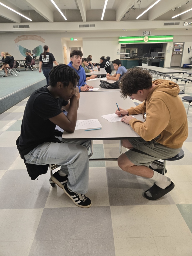 Two individuals are seated at a table in a cafeteria, engaged in conversation. One is writing, the other is listening.