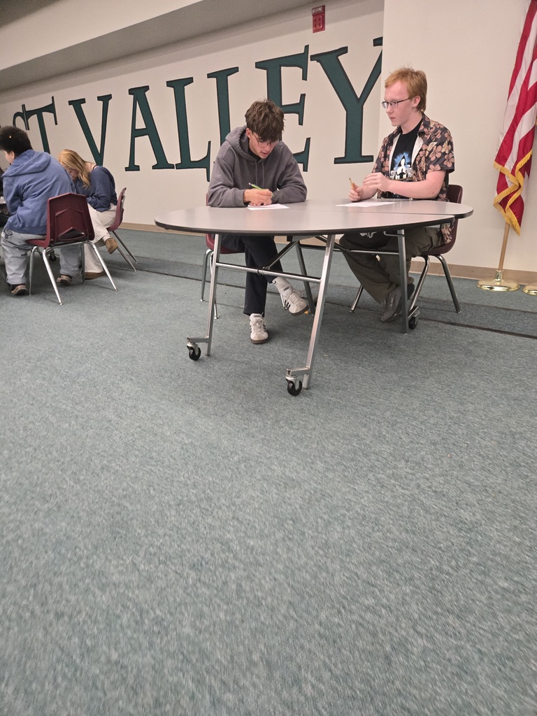 A room with people seated around a round table, writing. A US flag stands against the wall.