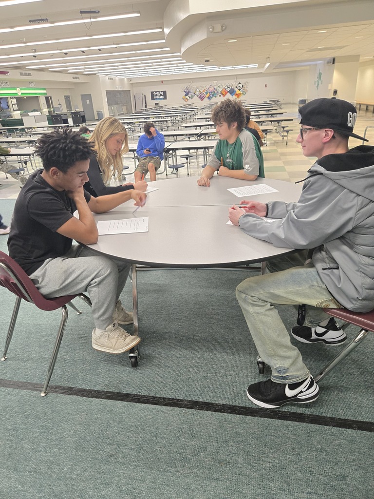 Four students sit around a round table in a cafeteria. Papers are on the table. The room has rows of tables and chairs.
