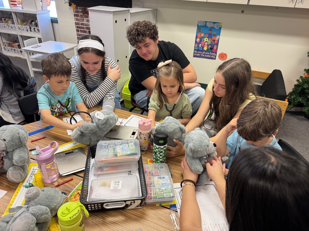 A group of children and adults gather around a table with stuffed toys, books, and a laptop.