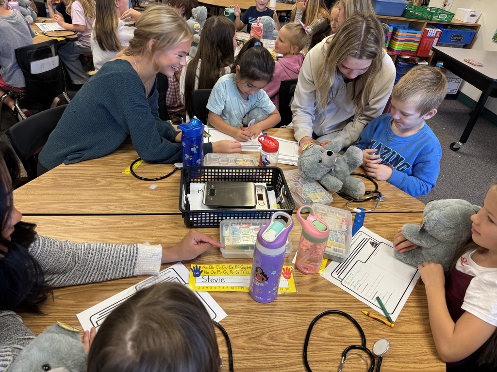 A group of children are sitting around a table in a classroom with adults, one holding a stuffed animal.