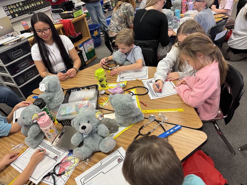 A classroom with young children and an adult. The children sit at desks, writing and drawing with stuffed toys and tools.