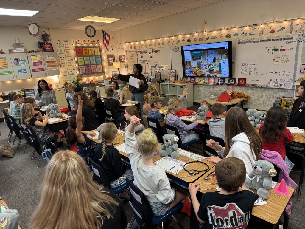 Classroom with students seated at desks, teacher at front, screen, some students raising hands, and plush toys.