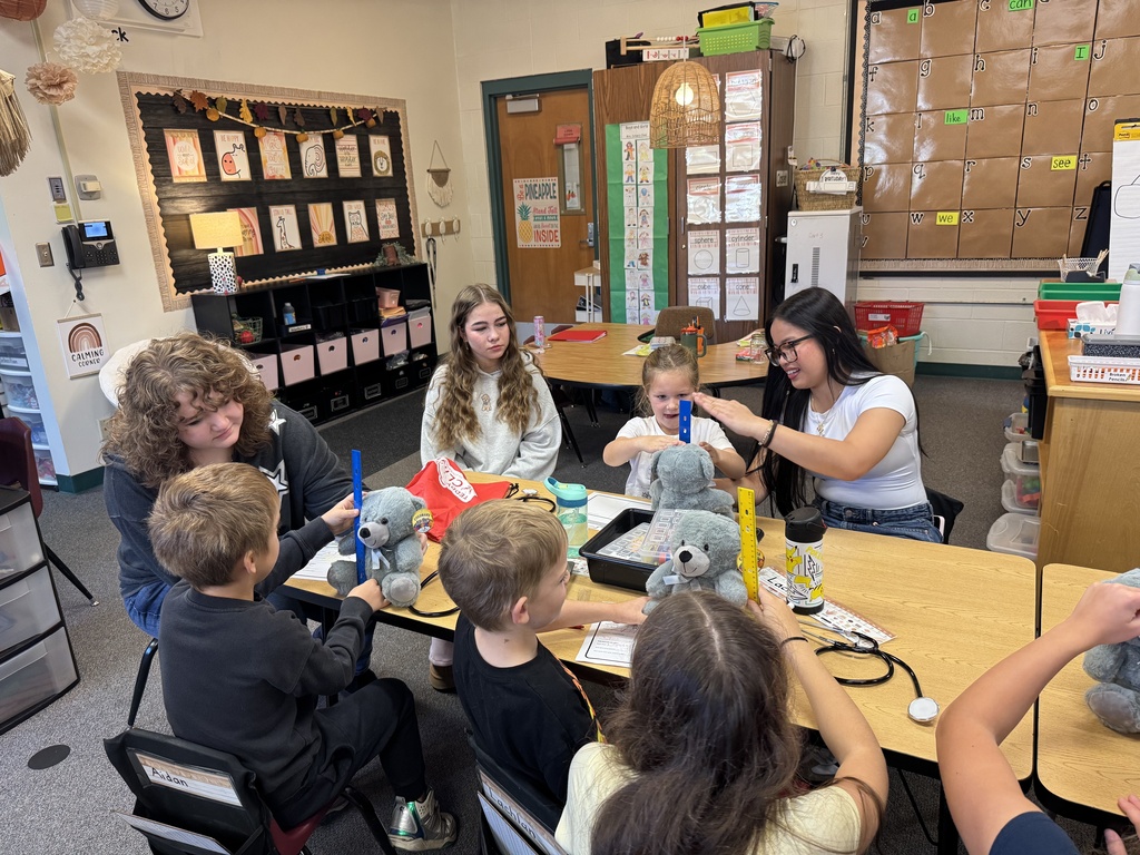 Several children are seated at a table, participating in a group activity with stuffed animals. A woman is standing nearby.