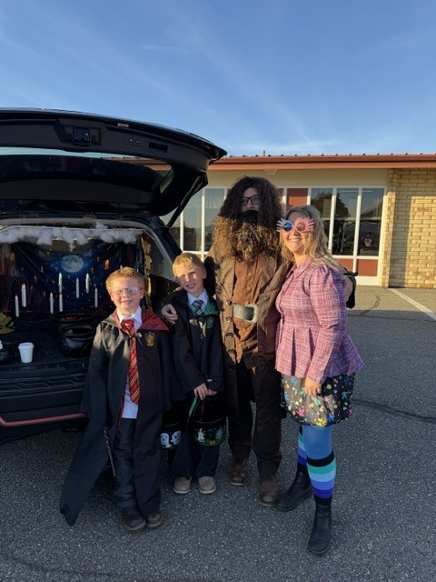 Four people in costumes pose by a car with its trunk open, in front of a building.