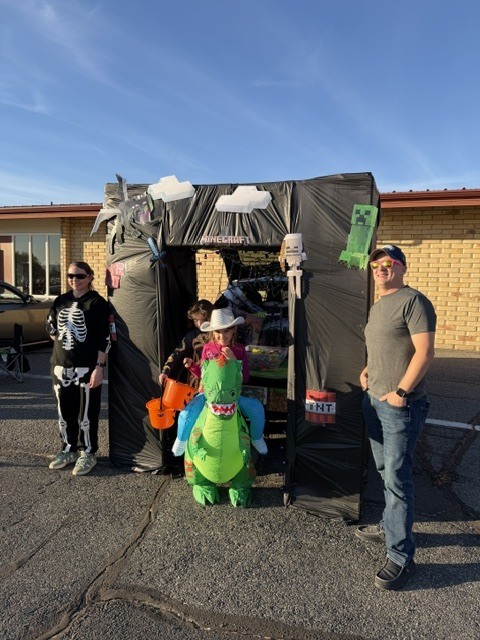 Four people in a parking lot, one wearing a hat and boots, one in a dinosaur costume, and one holding a bucket.