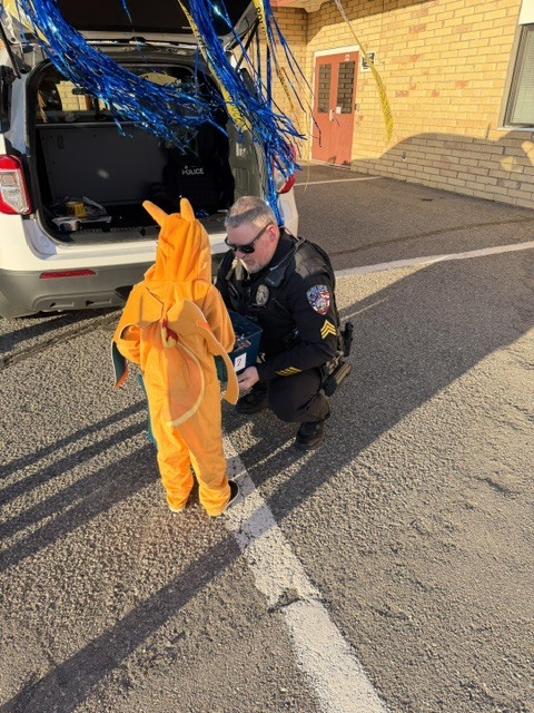 A police officer kneels with a young child in an animal costume, near an open car trunk.