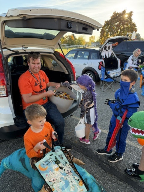 Adults and children in costumes, including a dragon, are gathered around a car with its trunk open.