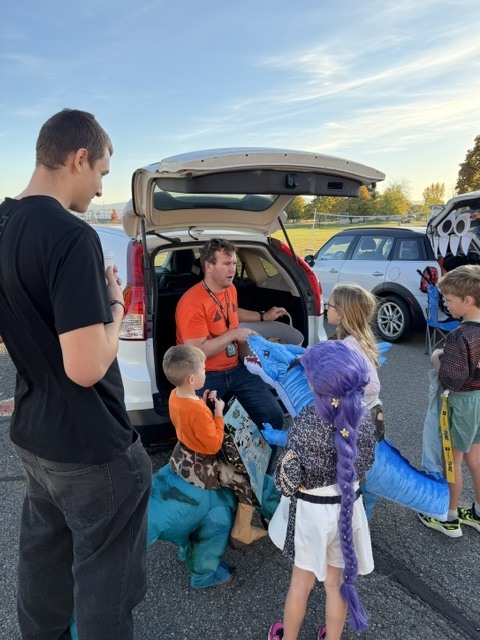 A group of kids and adults gather around a car with its trunk open. Kids wear costumes.