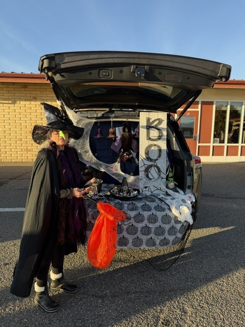 A person in a costume stands by a car with its trunk open. A table displays a sign.