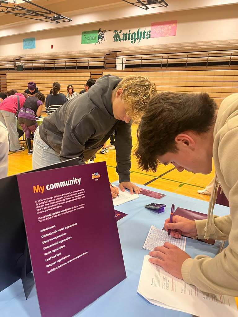 Two students at a table, one writing and the other reading, in a gymnasium with people gathered.