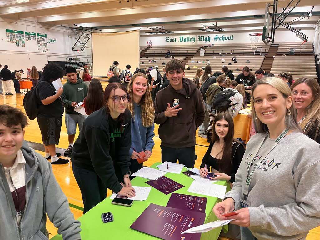 Students and adults gathered around a table with brochures and forms, standing in a school gym.