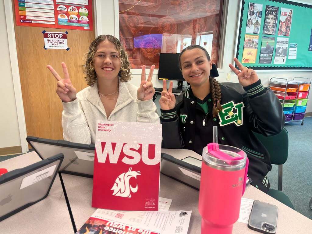 Two girls sitting at a table, making peace signs, with a pink tumbler and a WSU banner.
