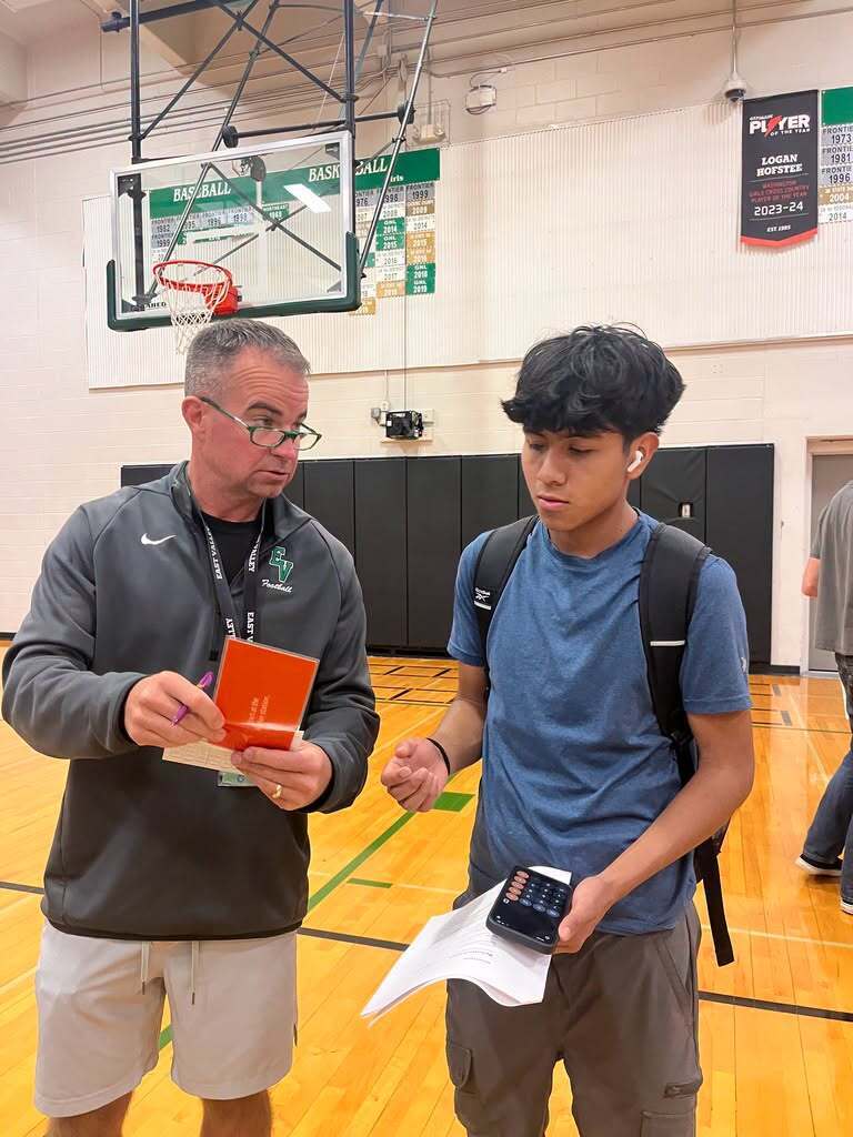 Two people in a basketball court. One man holds a folder, the other holds a device.