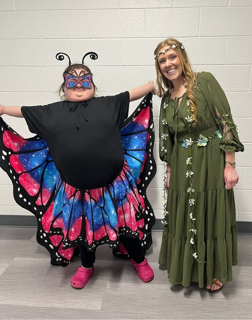 A smiling student displays her butterfly costume. She stands nexts to a staff member in a green woodland fairy costume. 