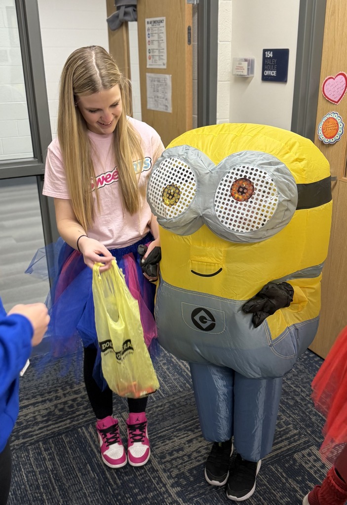 A smiling classroom parapro with a "Sweettarts" shirt and colorful tutu holds the hand of a student trick or treater in a minion costume. 