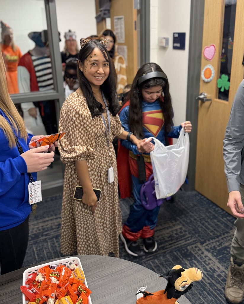 A smiling female classroom parapro holds the hand of a trick or treater dressed up as  Super Man. 