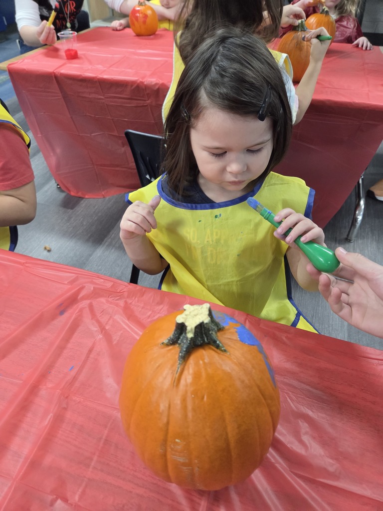 A preschooler in a yellow apron holds a paintbrush. A pumpkin with blue paint sits in front of her. 