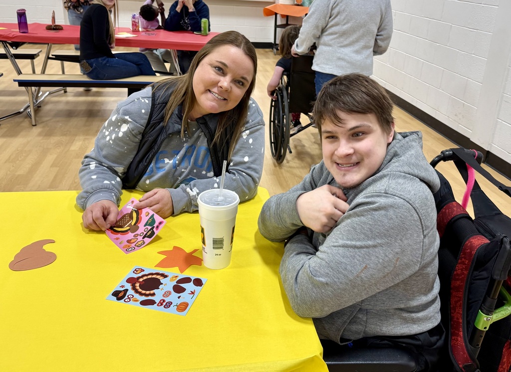 A smiling adult sits at a table with a smiling student in a wheelchair.
