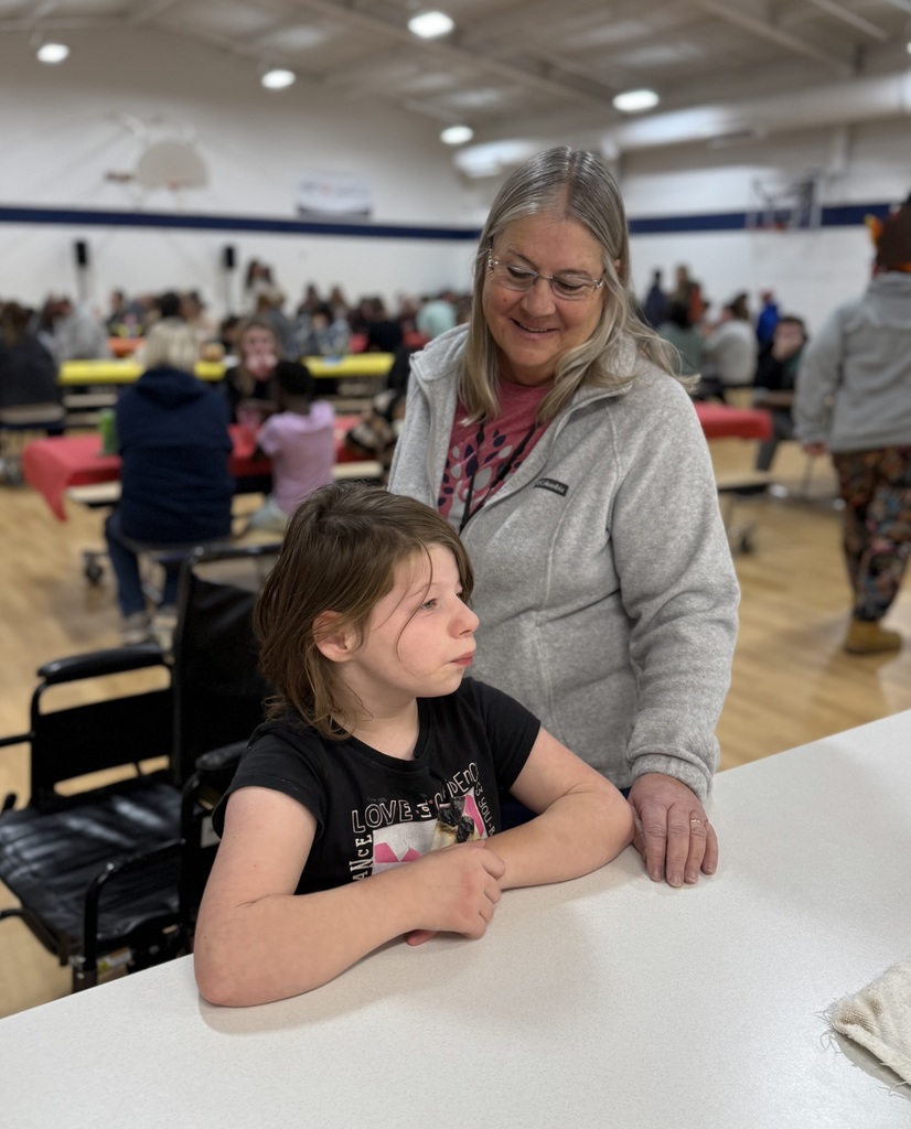 A young female student waits for the food to be served with her arms on the counter. An adult staff member stands behind her.