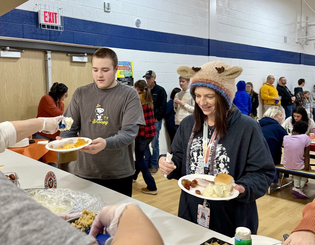 A student and staff member get their plates piled with Thanksgiving food. There is a line of people waiting their turn behind them.