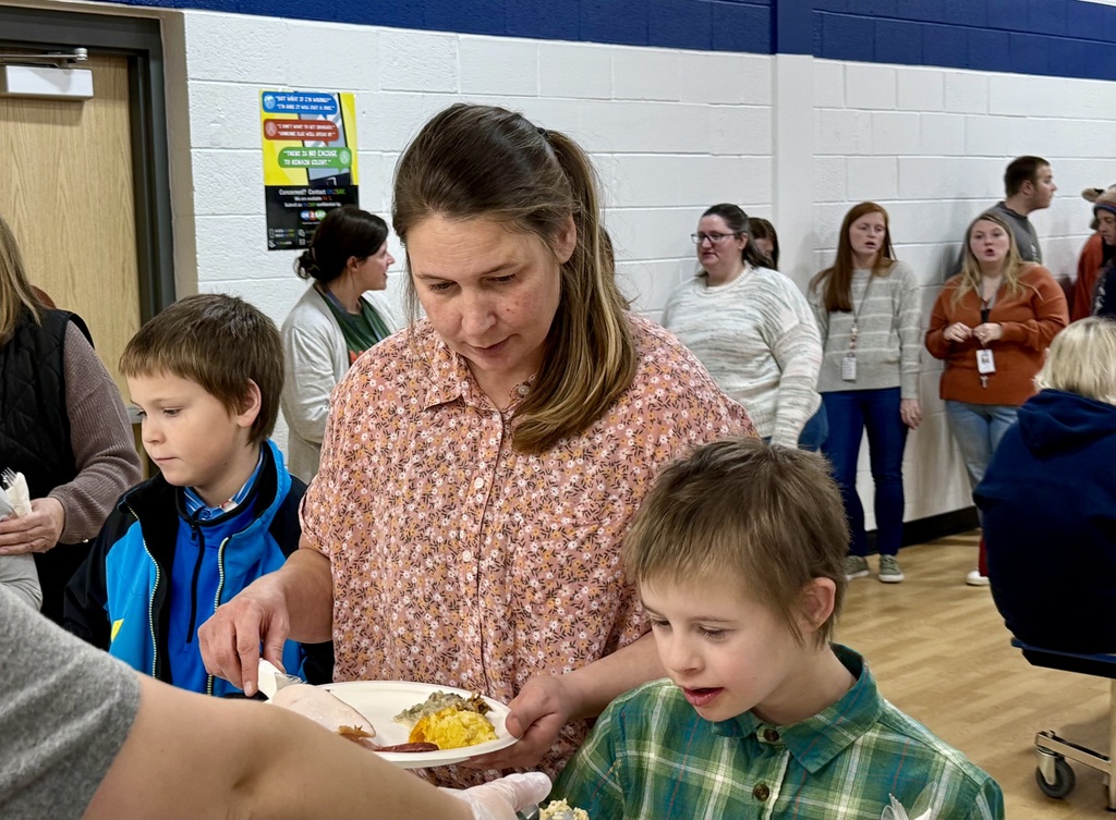 An adult with a student on each side of her get food in the serving line. A line of people can be seen behind them.