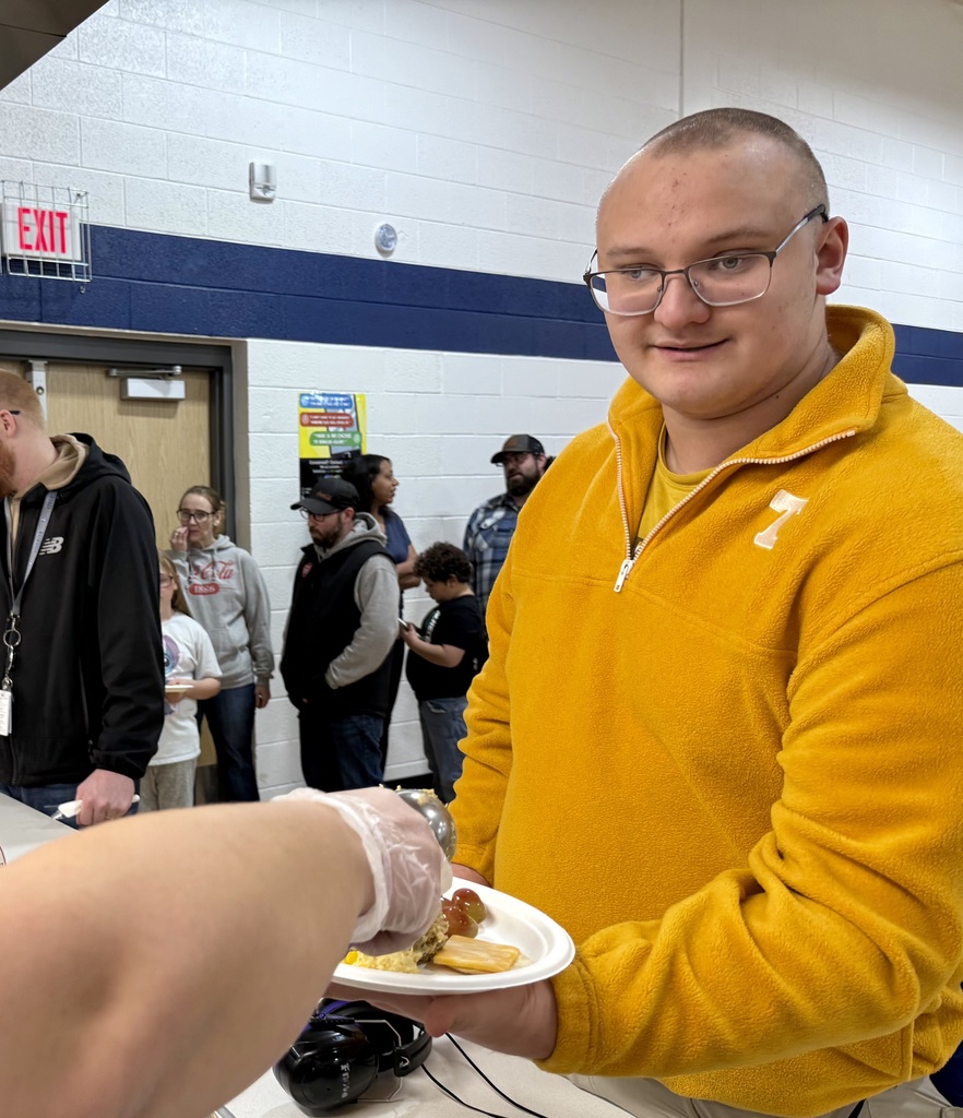 A smiling student holds his plate out. The arm of a staff member can be seen adding food to his plate in the serving line.