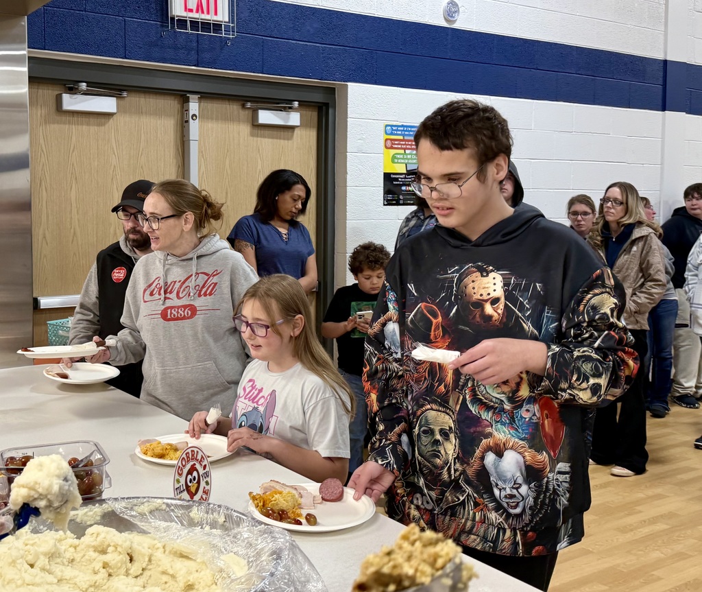 Smiling people carry plates through the food serving line. There is a line of people awaiting their turn in the background.