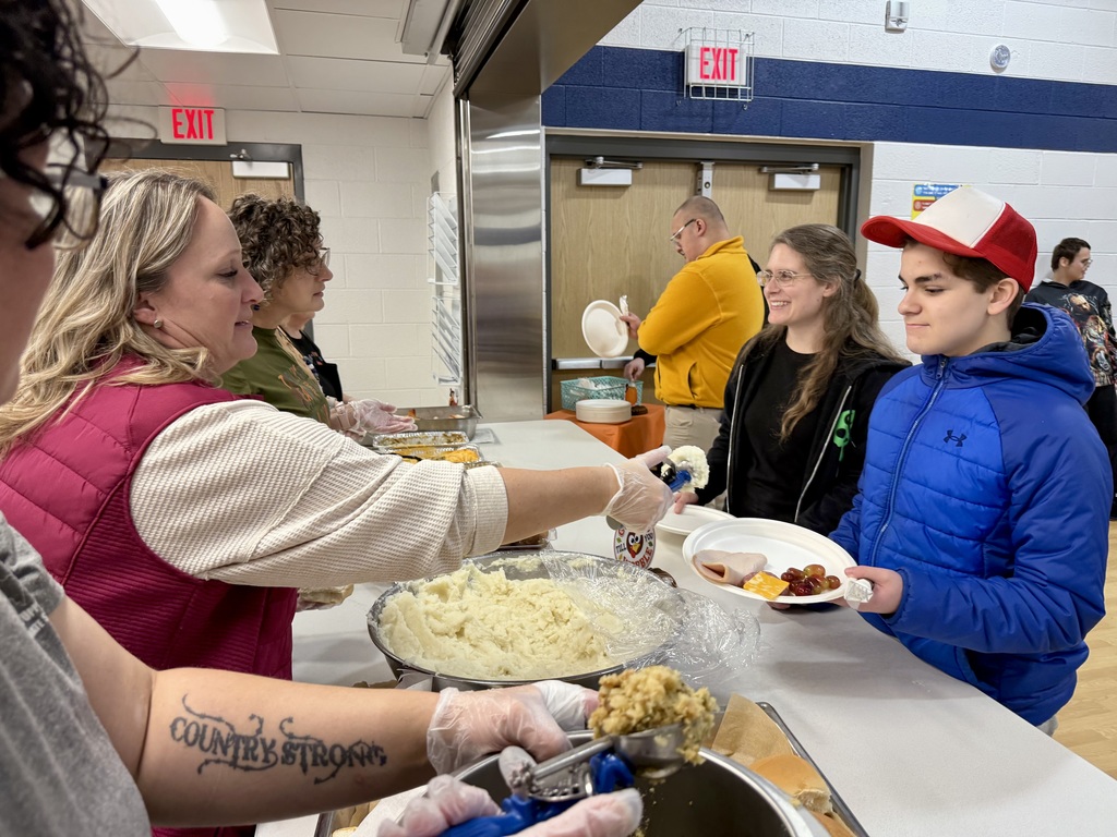 A smiling adult and a student carry their plates through the serving line, while staff can be seen scooping up mashed potatoes and stuffing.