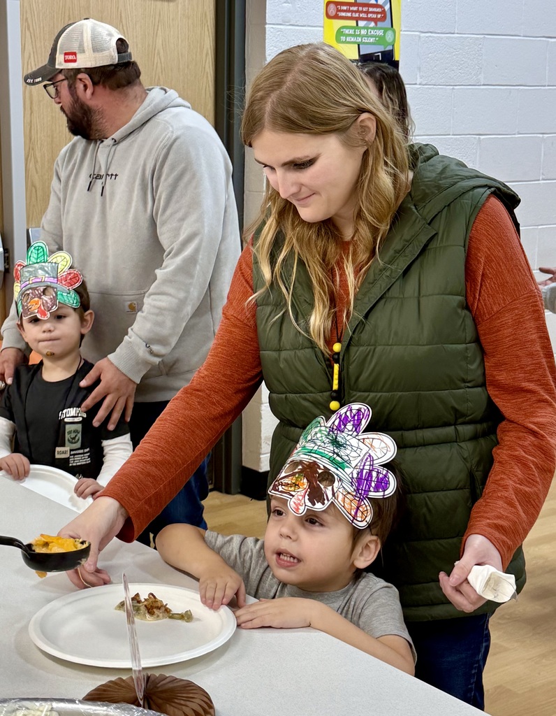 A staff member helps a young student who is peeking over the top of the counter and wearing a hand colored turkey hat.