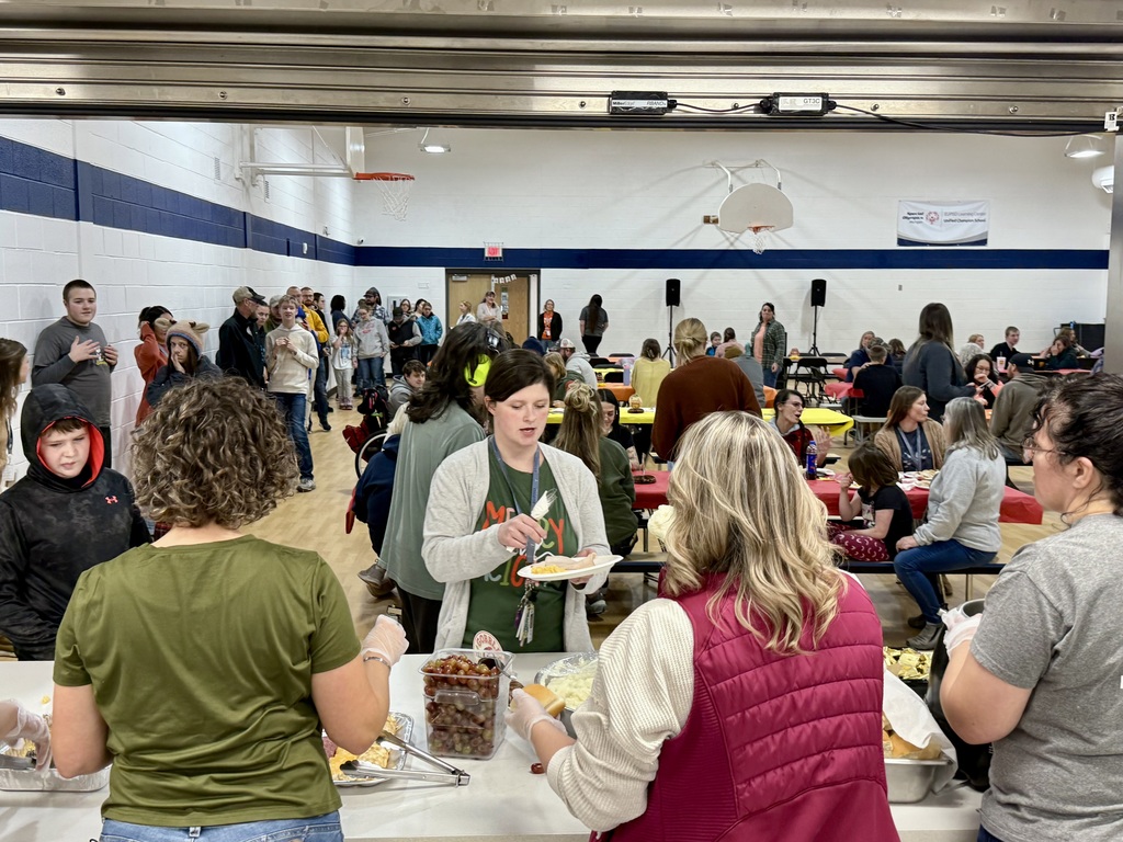 A view from behind the serving line of a gym full of people sitting at tables and a long line of people waiting for food along one side.