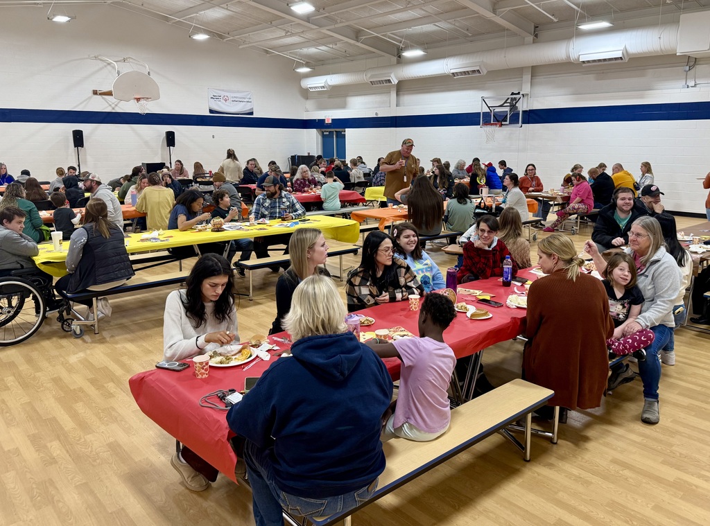 A view of the school gym filled with tables covered in bright yellow, orange and red tableclothes. People are sitting enjoying their Thanksgiving dinner.
