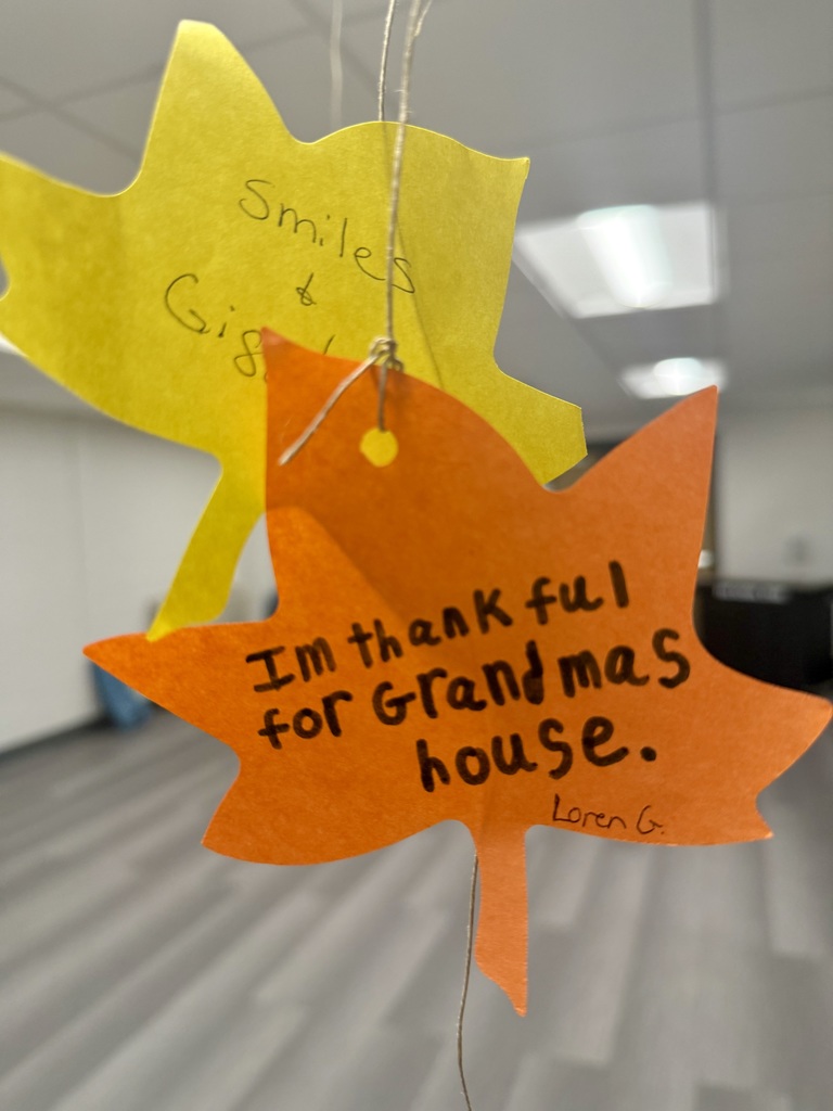 A bright orange construction paper leaf. The handwriting reads, "I'm thankful for Grandma's house." A yellow leaf hanging behind it, reads: "Smiles and Giggles."