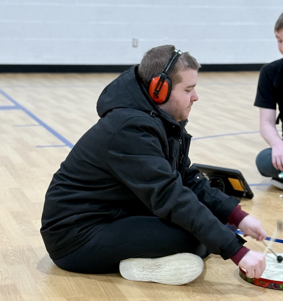 A student with headphones on sits on the floor with a small drum and two drum sticks. 