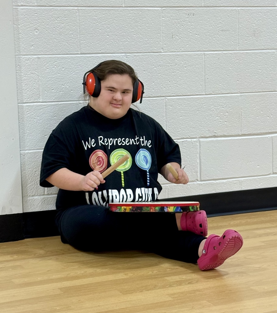 A student with headphones on sits on the floor with a small drum and two drum sticks. 