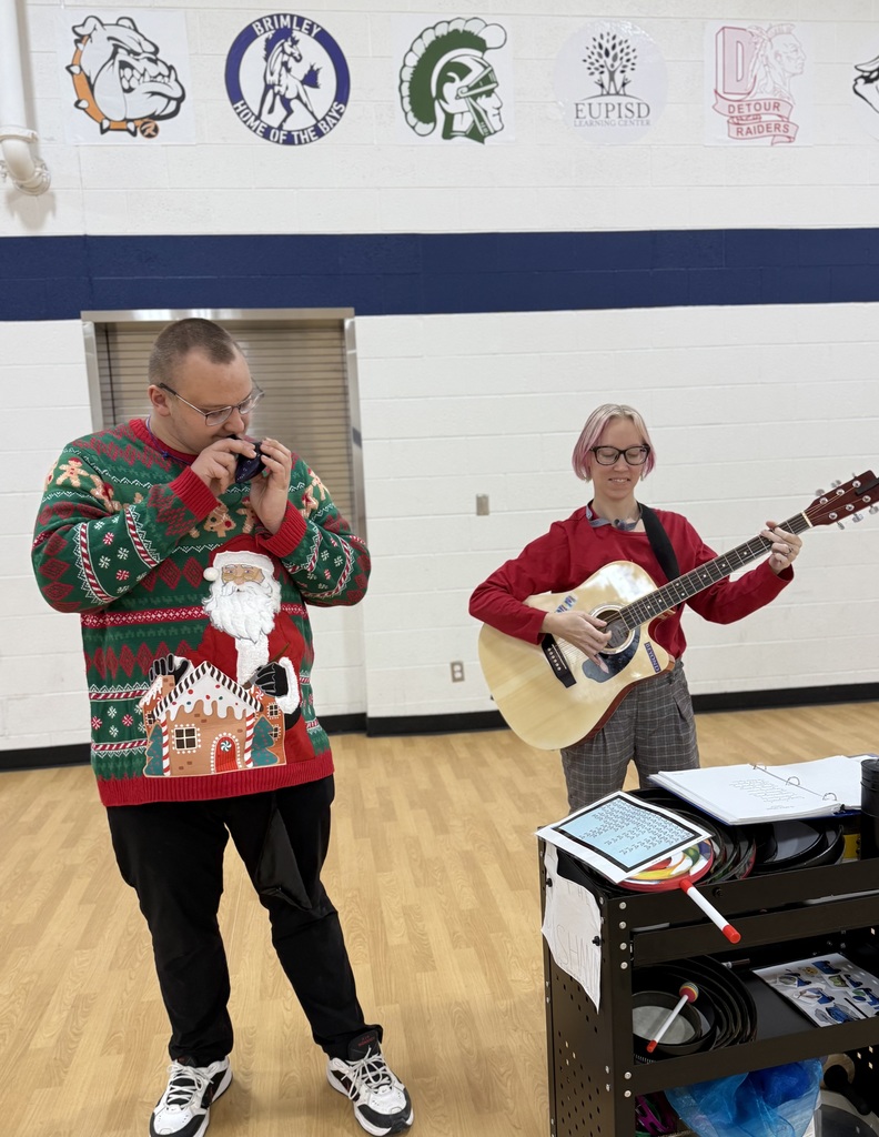 A smiling woman in a red sweater plays a guitar. a student in a vibrant holiday sweater stands nearby playing an instrument. 