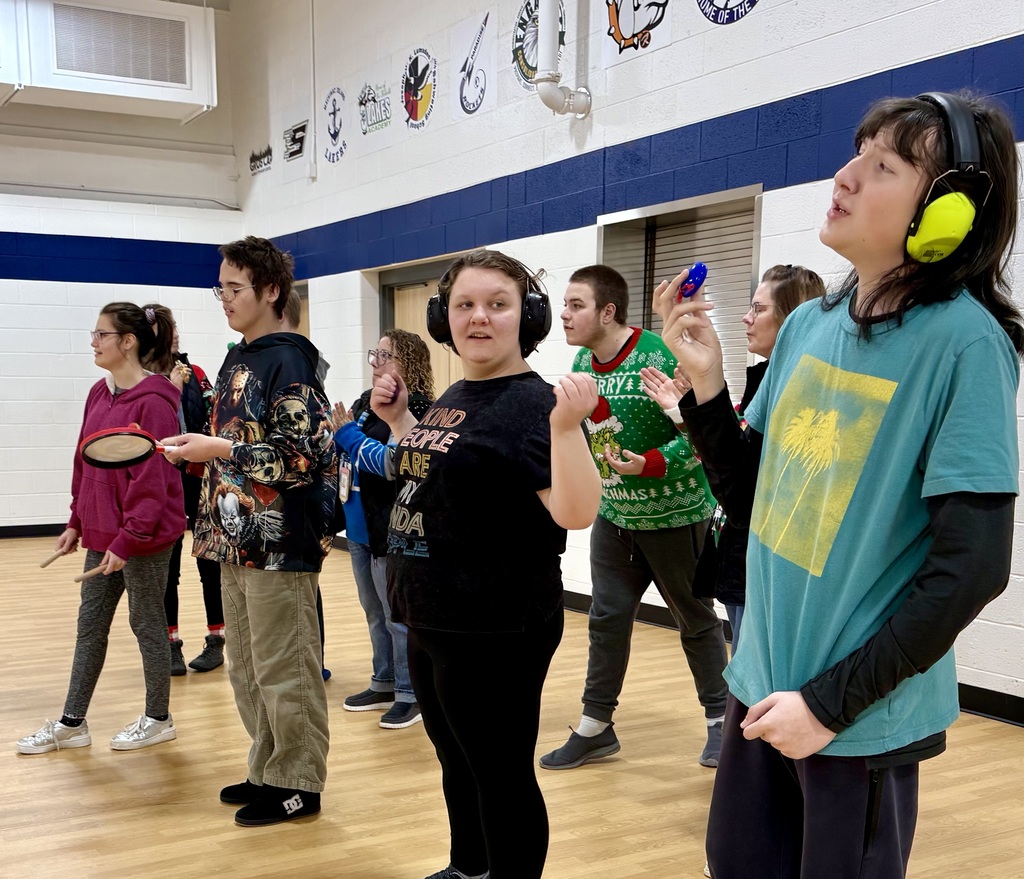 Students in holiday sweaters stand in two lines with hand held noise makers. They are in a school gym practicing  singing . 