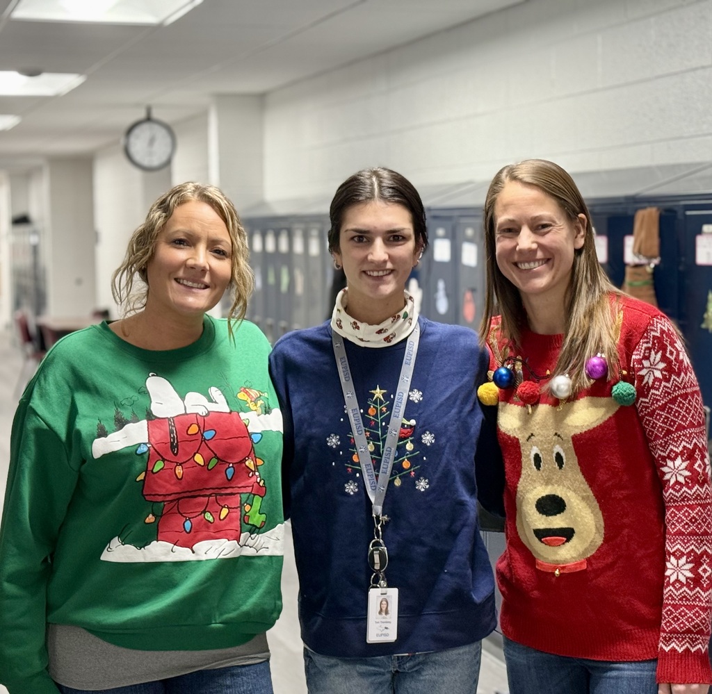 Three smiling staff member smile in their "ugly" holiday sweaters in a school hallway. There is a row of lockers behind them. 