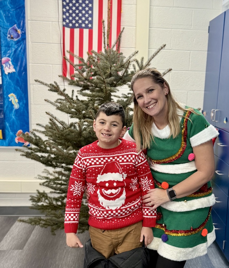 A smiling student in a bright red Santa Claus sweater poses in front of a  Christmas Tree with a staff member in a green and white striped,  sparkly sweater-dress. 