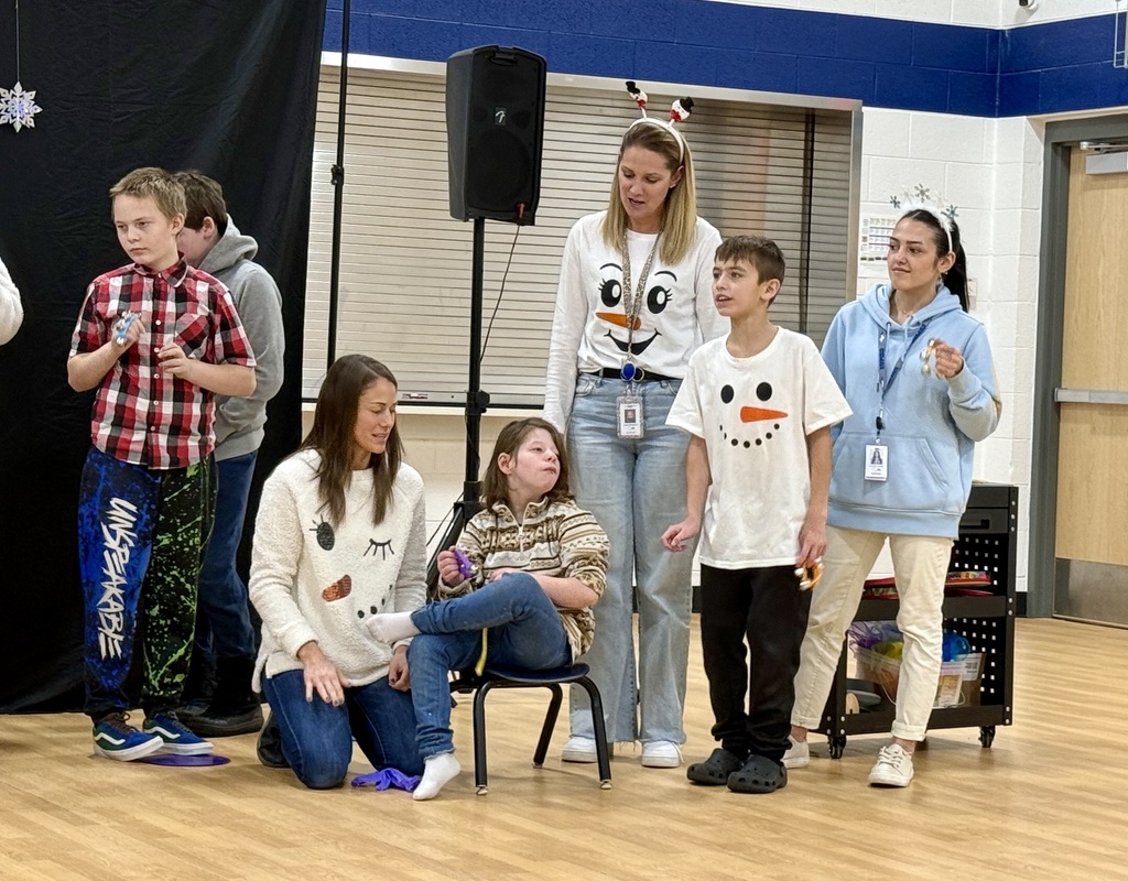 A young student sits in a chair with a teacher kneeling next to her. Another staff member and student stand on the other side of the chair. The two staff and student standing are all wearing snowman  shirts. 