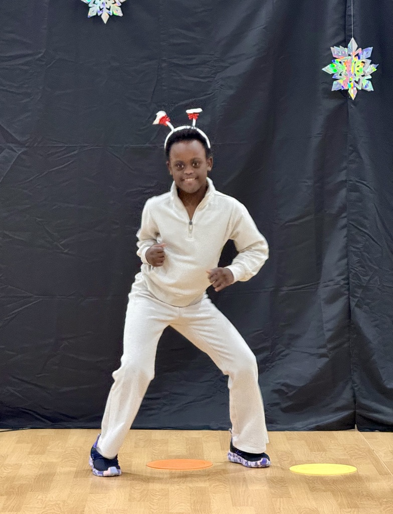 A young student in a white track suit and a holiday headband dances with a big smile on her face. 