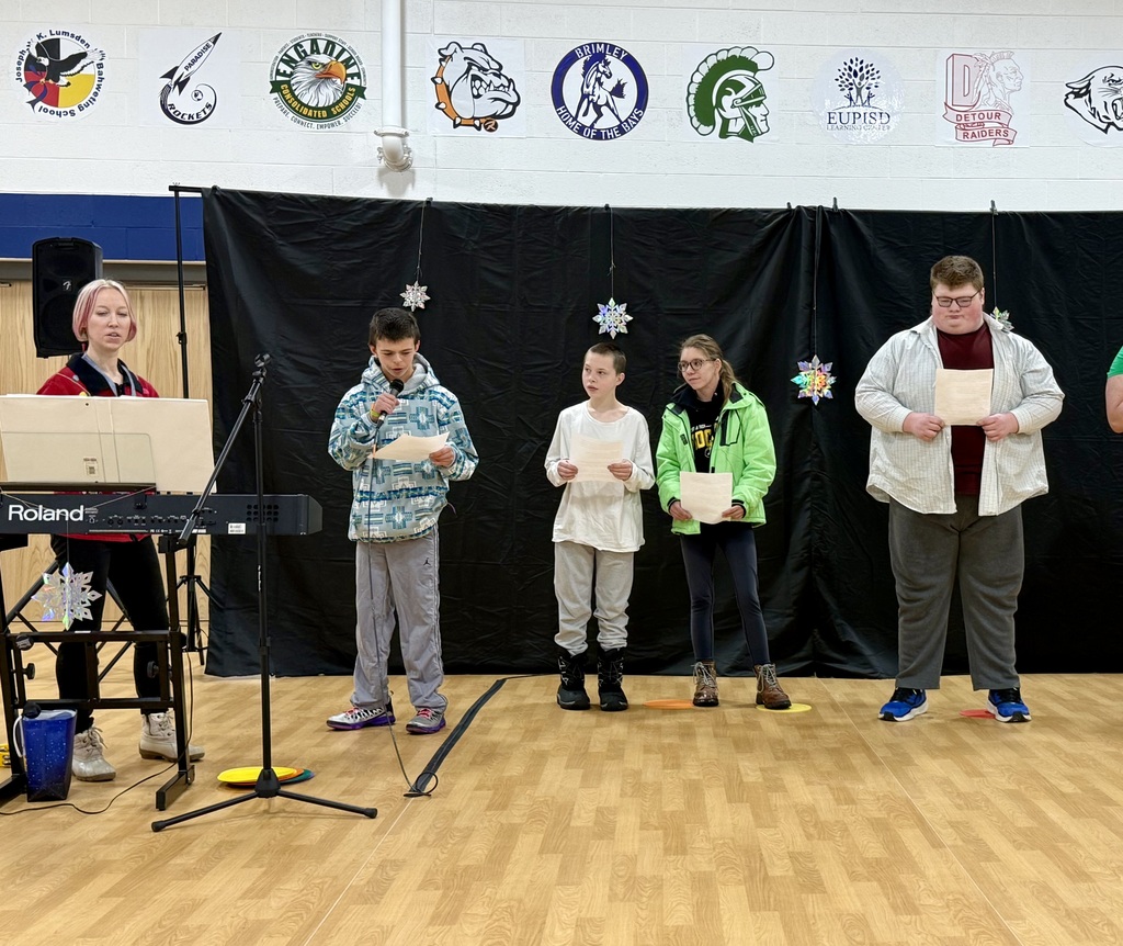 A line of 4 students stand with a sheet of song lyrics in their hands. One holds a microphone to speak to the audience. 